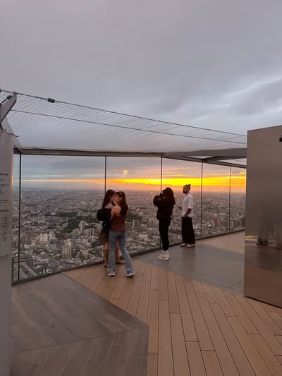 The image shows four people on an outdoor platform, presumably a high-rise observation deck, overlooking a cityscape at sunset. The location is Shibuya, Japan, recognizable by the urban layout and architecture. The platform has a wooden deck and a glass barrier with a protective net. The sunset paints the sky with hues of orange and yellow, visible through the glass and illuminating the cityscape below. The first person is in a white shirt and black pants. The second person is wearing a black jacket. The third and fourth people are standing closer, one in a brown top and jeans. They are facing outward, presumably looking at the sunset or the cityscape.