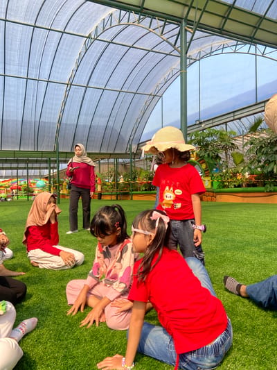 The image depicts a leisure area in Batu, Indonesia, characterized by a large arched canopy structure overhead and vibrant green artificial turf underfoot.

The scene features eight individuals, primarily children, and one adult woman. In the center-left, an adult woman stands upright, wearing a light beige hijab, a long-sleeved red top with cut-out shoulders, and dark trousers. She holds a smartphone in her right hand, her gaze directed towards it.

On the artificial turf, several children are seated. To the left of the standing woman, a girl wearing a light brown hijab, a red long-sleeved shirt, and light trousers sits cross-legged, with her left hand raised to her face. Adjacent to her, a second girl in a pink long-sleeved top adorned with cartoon characters and matching pink trousers sits cross-legged, her hands resting flat on the turf. In the right foreground, another girl in a red t-shirt and blue jeans is seated with her back partially turned, revealing a colorful bracelet on her left wrist.

Standing to the right of the center is a young girl wearing a large straw hat with a pink ribbon, light-colored sunglasses, a red t-shirt featuring a "CHANEL LABUBU" graphic, dark denim shorts, and a pink digital watch on her left wrist.

Partial views of other individuals are visible in the foreground: a person's lower legs in black trousers, another's legs wearing white socks with "GOOD DAY HAPPY" text, and a foot in a black and white striped sock and blue trousers.

The background reveals a recreational environment: colorful, mushroom-shaped playground structures and a rainbow arch are visible on the left, while dense green foliage and plants occupy the right. The overhead canopy structure consists of green metal beams forming an arched framework, supporting a translucent or semi-transparent roof material. An open section of the canopy structure reveals a clear blue sky. The lighting indicates bright daylight.