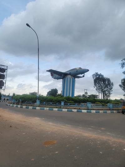 The image depicts a monument featuring a light blue jet airplane mounted on a blue and white striped cylindrical pedestal. The monument is situated in a traffic circle with an asphalt road. A white and blue striped curb surrounds a green landscaped area. In the background, a tall street lamp stands to the left, and trees are visible behind the monument. The sky is overcast with clouds. The monument's markings include the number "20" on the airplane's tail and "NAF" on its fuselage. The setting appears to be Barakin Lerawi, Nigeria, likely a public space or roundabout given the presence of the monument and road infrastructure.