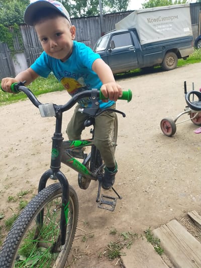 A young boy is captured in a close-up, slightly high-angle shot, sitting on a bicycle in an outdoor setting in Kuyeda, Russia. He wears a blue t-shirt with a "UNIVERSITY" print and a cartoon character, khaki pants, a grey and blue baseball cap, and black sneakers with green accents. The boy grins or smirks directly at the camera, holding the handlebars of his bicycle with bright green grips.
The bicycle has a black frame with vibrant green accents and green spokes on the front wheel. A white reflector is mounted on the handlebars, and a kickstand is visible on the left side, indicating the bike is stationary. The brand "STELS" is visible on the bike's frame, along with some numbers like "200."
In the background, to the right, a dark blue or green pickup truck with a light-colored canvas canopy is parked. The canopy features visible text in Cyrillic: "КомСикорм" (KomSikorm), along with two phone numbers: "8-908-257-05-26" and "8-912-059-15-69." A wooden fence and green foliage are visible further in the background. The ground is a dirt or gravel path with patches of grass and some wooden planks. Part of a red-wheeled tricycle is also visible on the right side of the frame. The lighting suggests it is daytime, possibly a cloudy day or in an area with indirect sunlight. The overall atmosphere appears casual and joyful.
