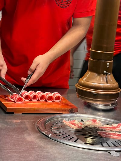 A person wearing a red shirt with a "YUKGA KOREAN BBQ" logo is preparing thinly sliced beef rolls on a wooden board. The beef rolls are arranged in a row. The person is using tongs and scissors to handle the meat. In the foreground, pieces of raw beef are cooking on a round metal grill. A large, copper-colored ventilation hood is positioned behind the grill. The scene appears to be set in a restaurant kitchen or dining area in Cebu City, Philippines.