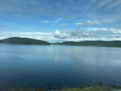 This image depicts a wide, calm body of water, likely a fjord or bay, with tree-covered hills and mountains forming the landscape on either side. The sky is a mix of blue and white clouds, with some cumulus formations indicating fair weather. The foreground shows a small strip of green grass and some dark rocks and vegetation along the water's edge. The scene is tranquil and natural, suggesting a location in a coastal region. Given the description of Skatval, Norway, this image likely captures a view of the Skatval Fjord or a similar coastal area within the municipality. There are no people or man-made objects prominently visible in the image.