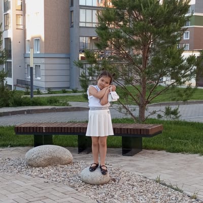 A young girl stands on a large, smooth stone in a courtyard, posing with her hands near her face and holding a small white purse. She is wearing a white sleeveless top and a white pleated skirt, with black Mary Jane shoes. Her dark hair is styled in two small buns on top of her head. Behind her, a modern wooden bench with dark metal legs is situated on a patch of green grass. The ground around the stones and bench is paved with interlocking gray bricks, interspersed with gravel. In the background, a tall, multi-story apartment building with a combination of brick and light-colored facades rises, with a mature pine tree in the mid-ground. The scene appears to be set during the daytime, under clear skies, as indicated by the bright sunlight casting shadows. The girl's pose and expression suggest a moment of playful pride or shyness. Text is visible on the waistband of the girl's skirt, but it is too small and pixelated to read. The location is Pervomaysky, Russia.
