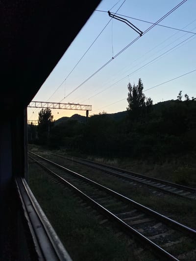 This media file captures a view of railroad tracks and overhead power lines in Mtskheta, Georgia. The scene is dominated by the railroad tracks, which run diagonally from the foreground into the distance, with multiple sets of parallel tracks visible. The tracks are situated alongside a grassy embankment and a dense, dark forest that climbs a hillside.
In the sky, numerous power lines crisscross the frame, supported by poles and insulators. A prominent metal lattice tower, characteristic of railway infrastructure, stands near the center of the image, with more power lines extending from it. The sky itself transitions from a pale, clear blue at the top to a lighter hue near the horizon, suggesting the presence of the sun just beyond the frame or set low in the sky. The lighting and the deep shadows cast by the trees and the foreground structure indicate it is either early morning or late evening.
The overall impression is one of a quiet, possibly rural or semi-urban, railway line at a time of day when natural light is soft. There is no visible text in the image. The composition, with the strong diagonal lines of the tracks and the silhouette of the foliage against the sky, creates a sense of depth and perspective. The dark foreground structure, possibly part of a bridge or overpass, frames the scene and adds to the dramatic contrast between light and shadow.