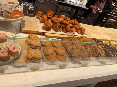 This image captures a detailed view of a bakery or cafe counter in Crawley, United Kingdom. In the foreground, a clear glass display case showcases various baked goods arranged on silver trays and a speckled white ceramic stand.
On the left, two small, round cakes with pink frosting and scattered pink garnish are displayed on the elevated stand. Adjacent to them, on a silver tray, stacks of "OAT, PECAN & CRANBERRY COOKIES" are visible, priced at £3.90. Moving right, another tray holds light brown, "RASPBERRY FILLED COOKIES," also priced at £3.90. Further to the right, a third tray presents "BARLEY CHOCOLATE & HAZELNUT COOKIES" and "CHOCOLATE CHUNK COOKIES" priced at £3.80, alongside other dark cookies with white toppings. Small wooden signs provide product names, prices, and calorie information for the items in the display.
Above the display case, in the midground, a pile of golden-brown "CINNAMON BUN" pastries, priced at £4.60, rests on a light wooden cutting board. Several other similar, empty wooden boards are arranged to the right, on a dark counter surface.
In the background, behind the counter, a person is visible wearing a dark jacket over a grey apron and a red lanyard with an ID badge. Their hands are holding a small white item. Behind them, dark industrial-style kitchen cabinets and appliances are present, including a "General Waste" bin on the left. To the far right, a tall, multi-tiered metal shelving unit is packed with numerous golden-brown baked items, likely bread or pastries.