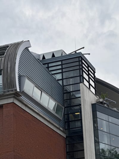 This is a medium shot, low-angle, close-up photograph of a modern building's exterior, featuring a mix of architectural styles and materials. The building's facade is composed of dark grey corrugated metal, red brick, and large glass panels framed with black metal. A distinctive curved section with a grey, metallic finish protrudes from the lower left of the frame. Above this, a steeply pitched section of the roof is covered in the same corrugated metal, with a narrow, horizontal skylight inset into it. Further up, a cantilevered section features extensive glass windows and a flat, light grey concrete surface. The sky above is overcast and grey, suggesting a cloudy or potentially rainy day. The lighting is diffuse, characteristic of an overcast sky. No people or specific activities are visible. The building's design is contemporary and geometric, with sharp angles and clean lines. The image appears to have been taken in a city, given the urban architectural style. There is no visible text in the image.