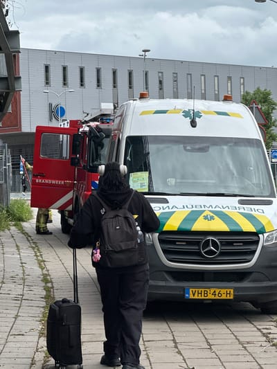 An image shows a person in black clothing, wearing headphones and carrying a black backpack and a black rolling suitcase, standing on a paved walkway in front of an ambulance and a red fire truck. The fire truck has "BRANDWEER" written on its side. The ambulance has a green and yellow stripe design and a star of life emblem. A person in a firefighter's uniform is visible near the open door of the fire truck. A building with a modern facade is in the background. A street sign with a bicycle symbol is visible to the right of the ambulance. The license plate of the ambulance reads "VHB-46-H". The scene appears to be a street in Amsterdam, Netherlands.