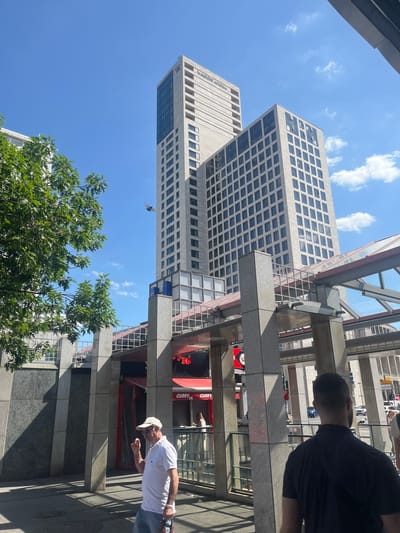 A man wearing a white polo shirt and a baseball cap stands on a sidewalk, holding a small object in his right hand. He is looking to his left. Behind him, a large, modern building with many windows dominates the background. A pedestrian walkway with a red railing runs across the mid-ground. A tree with green leaves is visible on the left side of the image. The sky is blue with scattered clouds. The scene appears to be in an urban environment. The text "CURRY 36" is visible on a red sign in the mid-ground, which is a well-known currywurst stand in Berlin, Germany.