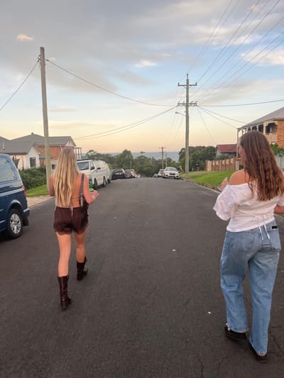 The image displays two females walking downhill on a paved residential street in Thirroul, Australia, during twilight. The sky features hues of orange, pink, and light blue with scattered clouds, and the ocean is visible in the distant background.

The female on the left, seen from behind, has long blonde hair. She is wearing a dark brown backless top, dark brown shorts with white lace trim, and dark brown mid-calf boots. She carries a dark shoulder bag and holds a green glass bottle in her left hand.

The female on the right, also viewed from behind, has long brown hair. She is wearing a white off-the-shoulder top with a tie-back detail, light blue wide-leg denim jeans, and dark slip-on shoes.

The street is lined with parked vehicles, including a blue panel van and a white panel van on the left side, and a white SUV further down on the right. Residential houses, constructed with varied materials including brick and light-colored facades, are situated on both sides of the street. Multiple wooden utility poles support numerous overhead power lines along the street. Green foliage, including trees and bushes, is present on the verges and in the distance.