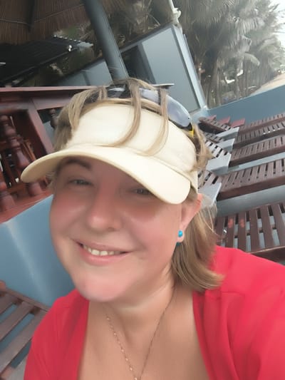 The media file is a selfie of a woman wearing a white visor and a red shirt. She has blonde hair and is smiling. She is wearing blue stud earrings and a delicate gold necklace. In the background, there is a thatched roof structure, palm trees, and what appear to be lounge chairs. The overall impression is that of a tropical vacation. The time of day appears to be daytime, and the weather seems to be overcast. The location context is Phu Thuy Ward, Vietnam.