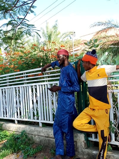 Two young men are standing by a white metal fence in Gombe, Nigeria. The man on the left wears a blue traditional outfit and a red kufi. He is smiling and holding a phone. The man on the right wears a yellow and black t-shirt and yellow pants. He has on an orange beanie with a blue and white pattern. He is looking to his right with his hand on his head. The background shows lush green foliage with orange flowers, palm trees, and a clear blue sky, suggesting a sunny day. The scene appears to be outdoors, possibly in a residential area or a park. There is some visible graffiti on the side of the pants of the man on the right. The overall mood seems casual and happy.