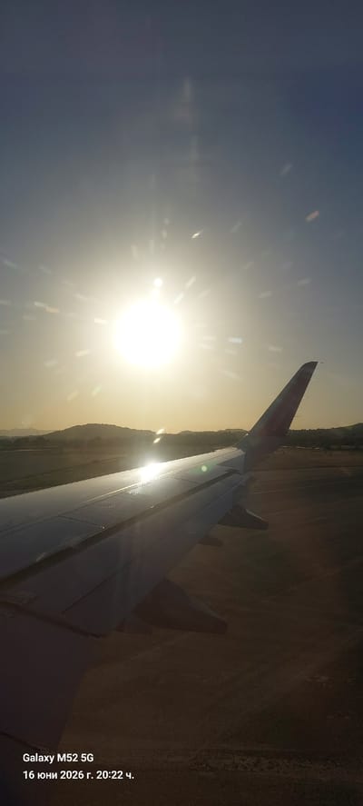 This is a vertical, close-up shot from inside an airplane, looking out over the right wing as the plane is either taking off or landing. The sun is low in the sky, casting a bright glare that creates lens flares and light streaks across the image. The sun's intense light illuminates the wing and the distant landscape of rolling hills. The wing itself appears to be a light gray color and is reflecting the sunlight. On the tail of the wing, the word "wizz" is visible in pink lettering, indicating the airline is Wizz Air. The background shows a flattened landscape with some vegetation and hills in the distance. The bottom left corner of the image contains text that reads "Galaxy M52 5G 16 юни 2026 г. 20:22 ч." This translates to "Galaxy M52 5G June 16, 2026, 20:22 h." The timestamp suggests this photo was taken in Manises, Spain, as the airport in Manises (Valencia Airport) is a major hub for Wizz Air and is served by this airline. The scene is dominated by the bright sunlight and the structure of the airplane wing.