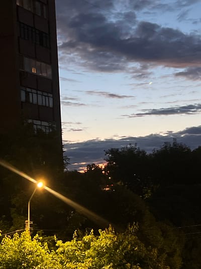The image depicts a nighttime scene in Pushkino, Russia, dominated by a tall, dark apartment building on the left. The building has multiple floors with visible windows, some illuminated from within. A street lamp is positioned in the foreground, emitting a bright yellow light that casts a strong glare across the lower portion of the image. Dense foliage, illuminated by the street lamp, is visible at the very bottom. Behind the building and trees, the sky is a gradient of twilight colors, with dark, cloudy formations against a lighter, pale blue sky. A sliver of a crescent moon is faintly visible in the upper right quadrant of the sky. The overall impression is that of an urban evening or night in a residential area.