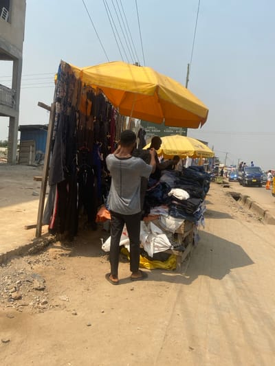 The image depicts an outdoor street vending scene in Ablekuma, Ghana, under clear, hazy daylight conditions. Two male individuals are positioned at a roadside market stall constructed from wooden frames and covered by two large, open yellow umbrellas. The individual in the foreground wears a light gray patterned t-shirt, dark trousers, and dark flip-flops, with his back partially to the camera, appearing to organize dark-colored apparel hanging on the left side of the stall. A second male, visible from the torso upwards, wears a dark shirt and stands beneath the second yellow umbrella, interacting with merchandise. The stall displays a variety of clothing items: dark trousers and tracksuits hang from the left side, while piles of folded blue jeans and other dark fabrics are stacked on a wooden pallet structure to the right. White plastic bags are present underneath the stacked clothing. A small sign behind the second vendor shows text including "WEDNESDAYS 9:00AM - 5:00PM" and "FRIDAYS 6:00PM - 8:00PM". The ground in the immediate foreground and under the stall is unpaved dirt and gravel. To the far left, an unfinished multi-story concrete building with exposed rebar is visible, alongside a small blue corrugated metal structure. A paved road with a concrete drainage ditch runs parallel to the stall on the right. Several vehicles, including a blue pickup truck, are present on the road in the background. Overhead utility wires traverse the clear, light blue sky. The sun is high, casting distinct shadows from the umbrellas and figures onto the ground.