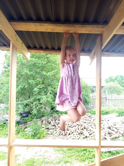 A young girl with blonde hair is hanging from a wooden beam with her hands. She is wearing a pink dress and patterned shorts. Her tongue is sticking out. The girl is in a semi-built wooden structure with a corrugated roof. In the background, there is dense green foliage, a pile of cut logs, and a wooden fence. The scene appears to be in a rural or suburban setting in Ostrovtsy, Russia, possibly a backyard or a construction site.
