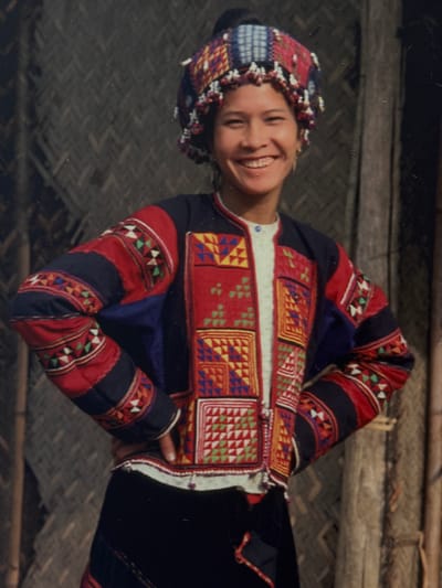 This is a vertically oriented, slightly blurry, color photograph of a young woman dressed in traditional clothing, likely from an ethnic minority group in Vietnam, given the location context of Hanoi. The woman is smiling broadly and looking towards the camera, her head tilted slightly. She is wearing an intricately embroidered jacket and a matching headdress. The jacket is predominantly red with black and navy blue accents, adorned with colorful geometric patterns in yellow, blue, green, and white, stitched in a patchwork style. The sleeves are also decorated with similar patterns. The headdress is a colorful band with beaded accents, possibly made of woven fabric. She is leaning on her arms, with her hands clasped in front of her chest. The background appears to be an interior setting, with wooden beams and woven mats, suggesting a rustic or traditional dwelling. The lighting appears to be natural, possibly daylight, but the exact time of day is not discernible. There is no visible text in the image. The overall mood is joyful and vibrant, highlighting the beauty of the traditional attire and the woman's cheerful expression.