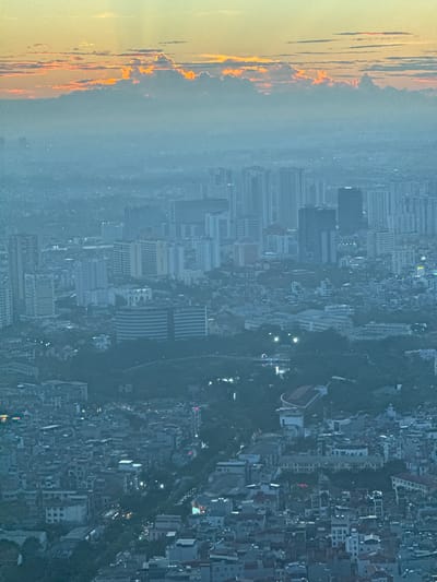 The image is an aerial view of Hanoi, Vietnam, likely captured at dusk or dawn. The sky is illuminated with warm tones of orange and yellow, with scattered clouds. The city below is shrouded in a blue-grey haze, typical of urban environments. A dense cluster of buildings, ranging in height from low-rise structures to taller, modern high-rises, fills the frame. Vegetation, in the form of trees and parks, is interspersed throughout the urban landscape. The lighting suggests that artificial lights from the city are beginning to illuminate, with some points of light visible in the distance. There are no visible people or animals in the image, but the presence of infrastructure indicates human habitation and activity. The overall impression is one of a vast, sprawling urban area under a dramatic sky.