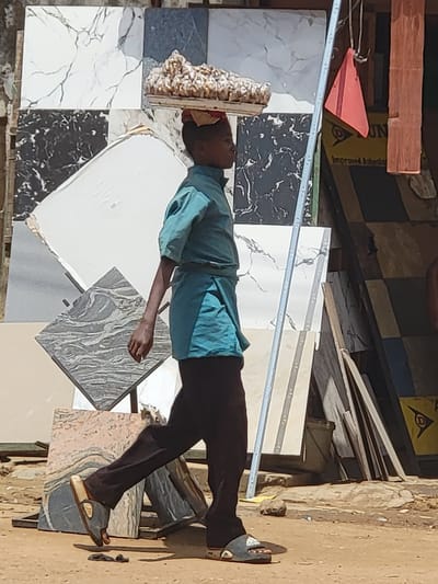 The image depicts an outdoor scene in Maiburiji, Nigeria, characterized by bright daylight. A young person, dressed in a blue-teal short-sleeved shirt, dark trousers, and sandals, is walking while expertly balancing a large, lumpy, light-colored load on their head. The person's face is partially obscured, but they appear focused on carrying their burden.
The immediate surroundings are dominated by a display or storage of various tiles, suggesting a tile shop, market stall, or construction material yard. These tiles come in an array of patterns and colors, including white with black veining, black with white veining, mottled grey, and a pinkish-beige marble-like design. Some tiles have visible silver-colored edge trims.
In the background, there are stacks of wooden planks and other construction debris. A yellow object or sign is partially visible, bearing the text "Improved Asbestos" and "DUNLOP," along with a triangular logo. The ground is unpaved, consisting of dirt or sand. The overall impression is one of a bustling commercial or work area under a clear, sunny sky.