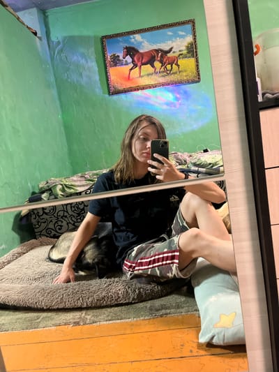A young woman with brown hair is taking a mirror selfie in a room with green walls. She is sitting on a large, fluffy dog bed with a dog lying next to her, partially under her arm. She is wearing a black t-shirt and camouflage shorts with red and white stripes. In the background, a framed picture of horses hangs on the wall, and there are green and blue lights projected onto the wall and ceiling. A mirror is positioned to capture the reflection, showing the woman, the dog, and the room. The floor in front of the dog bed is painted orange. A light blue pillow with a yellow star is visible to the right of the dog bed.
