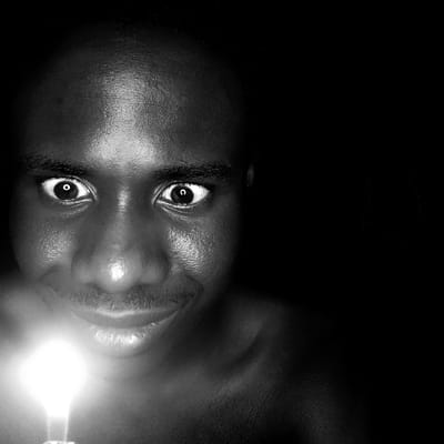 A black and white close-up image depicts a male individual, appearing to be of African descent, located in Narayi, Nigeria. The subject's face is illuminated from below by a single, bright light source, consistent with a lit candle, held out of frame below his chin. His eyes are wide open, pupils visible, and he exhibits a slight smile or smirk. Strong highlights are present on his forehead, nose, and cheekbones due to the upward light. The background is completely dark, suggesting an indoor setting at night or in a room devoid of other illumination. The individual appears bare-chested or wears dark attire that blends with the background.