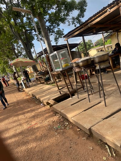 This media file captures an outdoor scene at what appears to be a roadside stall in Rafin Par, Nigeria. The image is oriented sideways. The primary subjects are several individuals and various objects associated with a food or vendor setup.
In the foreground, a person is seen walking on a dirt path, casting a long shadow, indicating it's likely late afternoon or early morning. Further into the scene, under a corrugated metal awning supported by wooden beams, are large metal pots, likely used for cooking. These pots are elevated on metal stands. Adjacent to this cooking area is a glass display case, possibly for showcasing food items.
Several people are visible in the background, some standing near the stall and others walking further down the path. One person is seated under a large, colorful umbrella, which is patterned with red stars and a logo that contains the word "Mar..." suggesting it's a branded umbrella. The surrounding environment shows trees with dense green foliage and a building in the distance. The ground is a mix of dirt and concrete, with some debris scattered around.
The overall impression is that of a busy commercial area, with vendors preparing or serving food, and customers or passersby. The lighting suggests a sunny day. There are no clear indicators of a specific event, but rather the daily operation of a roadside business. The mood appears to be one of everyday activity.
