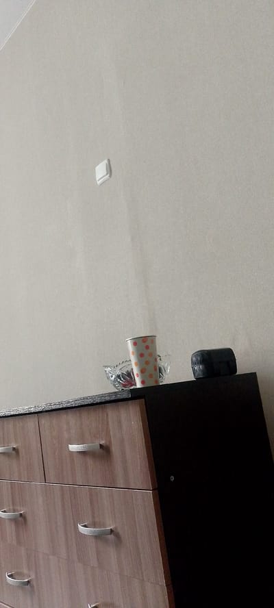 The image shows a dresser with several drawers and a dark side. On top of the dresser, there is a small glass bowl containing some items, a paper cup with an orange and pink polka dot pattern, and a black rectangular object. In the background, a plain, light-colored wall is visible, with a white light switch in the upper left quadrant. The dresser appears to be in a room, likely a bedroom or living area, given the presence of furniture and a light switch. The lighting suggests it could be daytime, but there are no clear indicators of the time of day or weather. There is no visible text in the image. The overall scene is still and domestic.