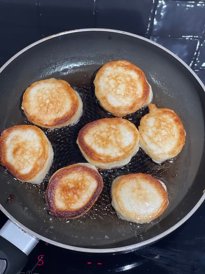In Zelenogradsk, Russia, seven small, golden-brown pancakes are being fried in a black, non-stick frying pan. The pan is placed on a black electric stovetop, with a red digital display showing the number "5". The pancakes are arranged in a circular pattern within the pan, with visible oil sizzling around them. The pancakes are plump and appear to be freshly made, with a light, airy texture visible on their surfaces. The stovetop has a silver handle on the left side, and the background is dark, with a slightly reflective surface that appears to be tiled.