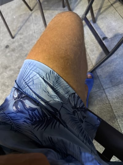 The image shows a close-up view of a person's legs and thighs, wearing blue patterned shorts. The person is wearing blue flip-flops on their right foot. The background consists of a tiled floor and the legs of what appear to be chairs. The lighting suggests an outdoor or well-lit indoor environment. There is no specific indication of the location being in Huangpu District, China, or any other specific city.