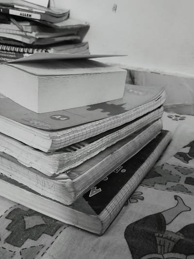 A stack of seven books lies on a patterned, textured surface. The top book is a thick, white paperback. The book below it is a grey paperback with the letters "A4" written on its cover. The remaining books have a similar, worn, textured look, though some are thinner. They are likely schoolbooks. The image was taken in the New Delhi, India area.
