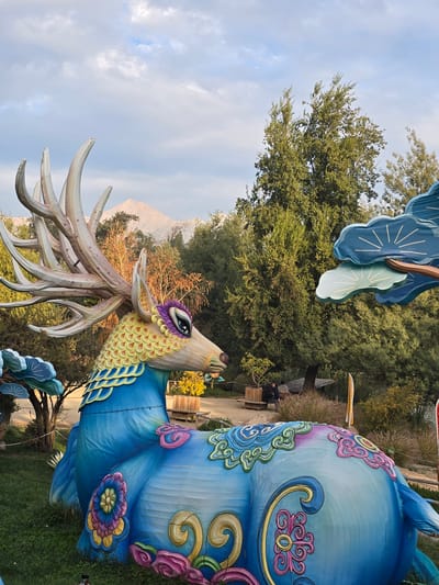A low-angle outdoor shot shows a blue and multicolored sculpture of a deer-like creature with large antlers. The sculpture is set in a park-like environment with trees and mountains in the background. In the background, there's a person sitting on a bench along a path. Based on the landscape features, this image was taken in Puente Alto, Chile.