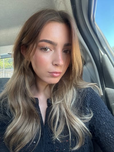 This image presents a close-up, head-and-shoulders portrait of a young adult female taken inside a vehicle in the city of Porto, Portugal.
The female subject has fair skin, brown eyes, and long, wavy hair that transitions from dark brown at the roots to lighter blonde or caramel highlights towards the ends. She wears light makeup, including defined eyebrows, mascara, a pinkish blush on her cheeks, and pink lip gloss. A small, light-colored stud earring is visible in her left ear. Her expression is neutral, and she looks directly at the camera. She is wearing a dark blue or black knitted sweater with a visible V-neck opening. The sweater has a coarse knit texture with scattered light-colored fibers or small flecks throughout its material.
The interior of the vehicle is partially visible, including a light gray headliner in the upper left corner and a dark gray or black door pillar and interior panel on the right side. Through the rear window on the left, an outdoor urban scene is visible under a clear, blue sky. This background includes green tree foliage behind a light-colored stone or concrete balustrade or railing, indicating an elevated position or a street-side view. No specific identifying landmarks within Porto are discernible from these background elements. The subject is engaged in self-portrait photography.