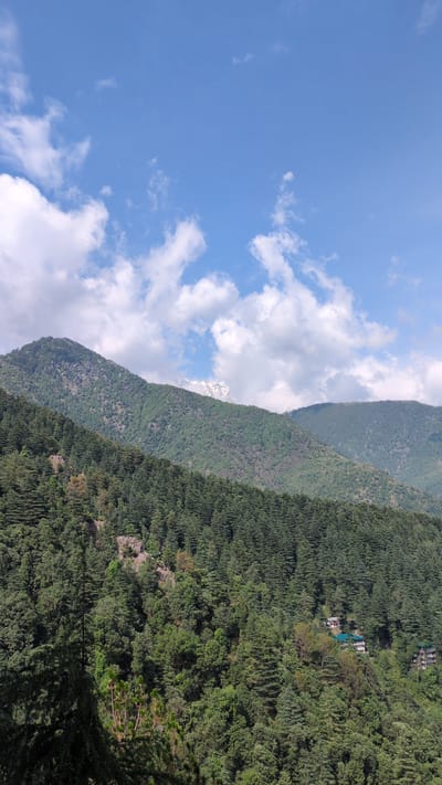 The image depicts a mountainous landscape in Dharamkot, India. The sky is blue with scattered white clouds. Two mountain ridges are visible, covered in dense green vegetation. On the right-hand side, a cluster of buildings is nestled amongst the trees on the mountainside. The mountains appear to be rugged and densely forested. In between the two mountain ridges, a glimpse of a snow-capped peak is visible through a gap in the clouds.