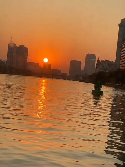 The image depicts a serene sunset or sunrise over a city, likely Guang Zhou Shi, China, with a prominent body of water in the foreground. The sky is bathed in warm, intense orange and yellow hues, indicative of the golden hour.

The sun, a bright white orb, is positioned low on the horizon, partially obscured by the silhouettes of several urban buildings. Its powerful light casts a warm, hazy glow across the entire atmosphere. The city skyline, consisting of numerous high-rise buildings and structures, stands against the bright sky, appearing as dark, misty shapes on the left side of the frame.

The right side of the image is dominated by the calm surface of the water, which beautifully reflects the sun's brilliant light. A shimmering, vertical column of bright orange and yellow light extends across the water from the sun, creating a striking focal point. The water's surface shows subtle ripples and textures, enhancing the reflective patterns.

In the middle ground, a distinctive green buoy or navigational marker floats on the water, providing a contrast to the warm color palette of the scene. The overall impression is one of peaceful urban beauty, with natural light interacting dramatically with the city and water. There is no visible text in the image.