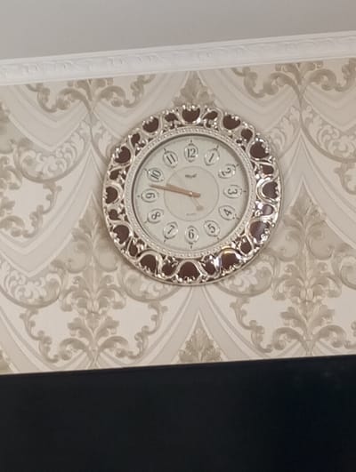 The image displays an interior scene from a room in the city of Яромаска, Russia. A circular wall clock is centrally mounted on a wall covered with patterned wallpaper. The clock features an ornate, silver-colored frame with dark reddish-brown accents, surrounding a white face with Arabic numerals (1-12). Gold-colored hour and minute hands are set to approximately 8:50 or 9:50, with the minute hand pointing towards the "10" and the hour hand positioned between "8" and "9". The brand "Mepoli" and "Quartz" are visible on the clock face. The wallpaper exhibits a repeating damask pattern in shades of light beige and grey. Above the wallpaper, a white, decorative crown molding separates the wall from a plain white ceiling. The lower portion of the image is occupied by a large, dark, featureless rectangular object, possibly a television screen or a piece of furniture. No individuals, actions, or interactions are visible.