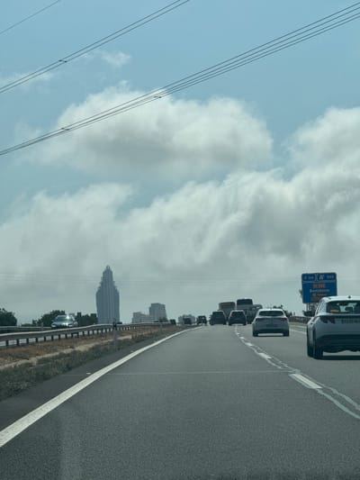 The image depicts a view from a vehicle traveling on a multi-lane highway in the vicinity of Villajoyosa, Spain. In the distance, a distinctive skyscraper with a stepped, tapering profile is visible, likely indicating the Benidorm skyline. The highway has clear lane markings and a metal guardrail runs along the side, separating it from a dry, scrubby verge. Several vehicles, including cars and trucks, are on the highway, suggesting moderate traffic. A blue highway sign is visible on the right, indicating "Benidorm" and distances to towns. The sky is partly cloudy, with large, white cumulus clouds against a pale blue backdrop. Overhead, multiple power lines are strung across the sky. The overall scene is one of transit and urban sprawl.