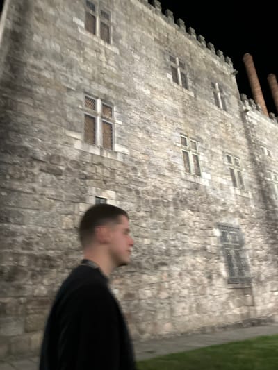 An outdoor, nighttime photograph depicts the side of the Palace of the Dukes of Braganza (Paço dos Duques de Bragança) in Guimarães, Portugal. The building features a facade of light grey, rough-hewn stone blocks, illuminated externally. Multiple rectangular windows, each with stone mullions and transoms forming a cross pattern, are visible across the wall. Several windows include metal grates or bars externally. The roofline is crenellated, and two tall, cylindrical, reddish-brown brick chimneys rise prominently on the upper right side against a dark night sky. In the foreground, on the left, a male individual is captured in profile, facing right. He possesses short, dark hair and wears a dark, long-sleeved garment with a distinct patterned collar or neckline visible. The individual's features are slightly blurred, indicative of motion or shallow depth of field. A narrow strip of green grass and a paved surface are visible at the base of the building on the lower right.