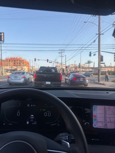 The image, taken from the perspective of a driver inside a car, shows a view through the windshield at an intersection in Los Angeles, California. The time is 7:05 AM. The sky is clear and blue. Several cars are stopped at the red traffic lights. A silver sedan is in the left lane, a black pickup truck is in the center lane, and a dark purple sports car is in the right lane. In the background, there is a multi-story building under construction. Traffic lights and utility poles with numerous wires are visible throughout the scene. The car's dashboard displays the current speed as 0 MPH, the gear as 'D', and the outdoor temperature as 87°F. The infotainment screen shows a navigation map and the current time. A person is visible on the sidewalk to the right of the intersection, holding a blue bag.