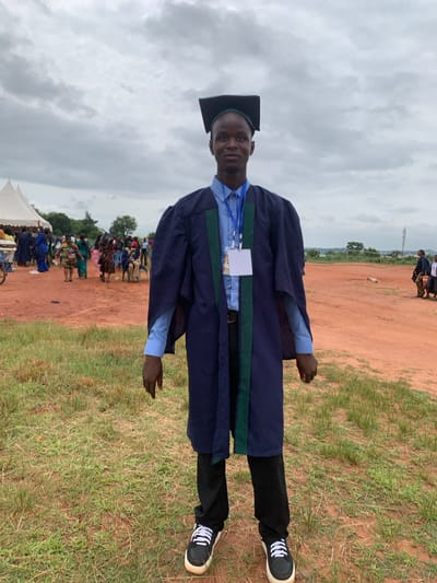 A young man in a graduation gown and cap stands in a field, likely in Nsugbe, Nigeria. He wears black pants and black and white sneakers. There are people gathered in the background. The ground is a mix of red dirt and grass, with an overcast sky above. A building or a hill can be observed in the distance.