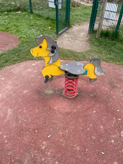 This image captures an outdoor playground scene in Holtzheim, France. A yellow and gray horse-shaped spring rider, equipped with black handles and footrests, is centrally positioned. It is mounted on a red coiled spring anchored into a reddish-brown, worn, and cracked rubberized safety surface. This surface exhibits sparse small white debris and one visible cigarette butt. Surrounding the safety surface is a green grassy area containing scattered dry leaves. In the background, a green metal fence with an open gate leads to a dirt path extending into another vegetated section. A wooden post stands to the right of the open gate. The scene is devoid of human presence or activity.