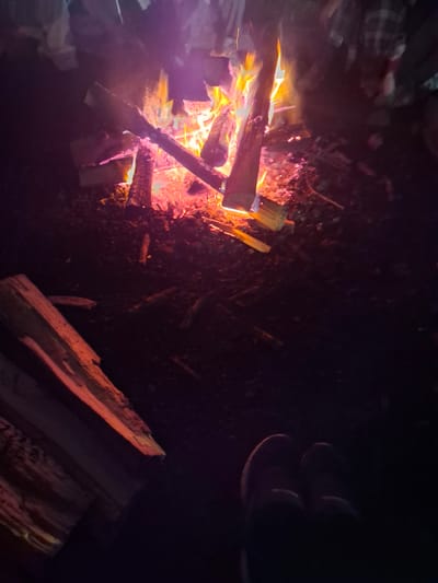 This is a nighttime photograph of a campfire. The scene is dark, with the fire being the primary light source, casting a warm orange and pink glow. Several logs are burning, with visible flames licking at them. Some unburnt logs are stacked nearby, also illuminated by the fire.

In the upper left corner, partially visible, are what appear to be people sitting around the fire. One person is wearing a plaid shirt, suggesting casual attire suitable for an outdoor gathering. The people are largely out of focus and silhouetted against the darkness.

To the right of the campfire, a pair of dark shoes or sandals are visible, also resting on the ground. The ground appears to be dirt or a similar natural surface, with scattered leaves or debris.

The overall mood is one of a cozy and relaxed evening, typical of a campfire gathering. The time of day is clearly night. There are no explicit weather cues, but the presence of a campfire suggests it is likely cool enough for one. The location cues are minimal, but the setting appears to be outdoors, possibly a backyard, campsite, or rural area. No text is visible in the image.