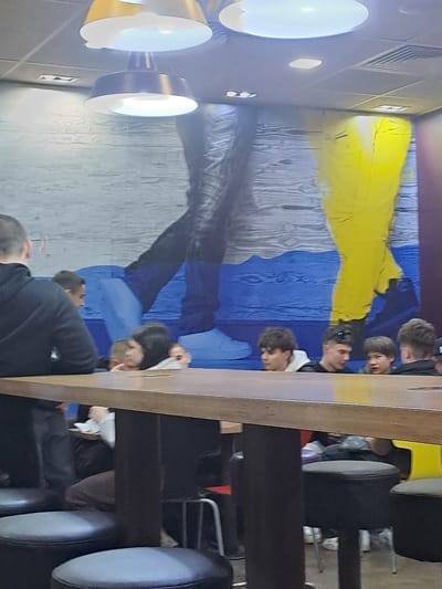 The image shows the interior of a dining establishment, possibly a fast-food restaurant. A group of people are seated at a long table, most of whom appear to be teenagers or young adults. They are positioned against a wall mural depicting the flag of Ukraine. The furniture consists of high stools with dark upholstery and metal frames. Overhead lighting fixtures are visible. The location is identified as Elektrostal, Russia.