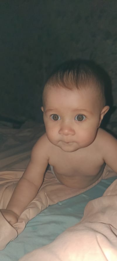 A close-up shot captures a baby crawling on a bed. The baby, with light brown hair and wide, alert eyes, is positioned in the center of the frame, looking directly at the camera. Their skin appears fair, and they are unclothed from the waist up, with a diaper visible beneath. The bed is covered with light pink and teal linens, which are slightly wrinkled, suggesting a lived-in space. The background is dark and out of focus, with a hint of a wall or backdrop. There is no indication of the location being in the city of Хохлы, Russia, nor any specific environmental context beyond the bedroom setting.