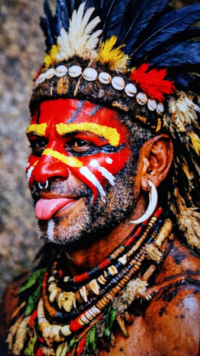 The image is a close-up portrait of a man with his face painted in red, yellow, and white patterns. He is wearing a headdress adorned with feathers and shells, and several necklaces made of beads, shells, and what appear to be bone fragments. He has a septum ring and a large, curved earring made of a bone or tusk. His tongue is extended slightly from his mouth. The background is a blurred, textured surface. There is no indication of the city of Riga, Latvia, in the image.
