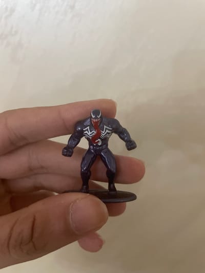 A human hand with light-to-medium skin tone holds a small, dark blue or purple Venom action figure against a plain, light beige background. The figure, approximately 2-3 inches tall, stands on a small circular base and is positioned towards the center-right of the frame, with the hand partially visible on the left and bottom, out of primary focus. The Venom figure exhibits its iconic white spider symbol on the chest, white eyes, an open mouth with sharp white teeth, and a long, red tongue protruding forward. Its body is muscular, and its fists are clenched, conveying a dynamic pose. The background wall is uniform in color, showing minimal texture or detail. The scene provides no visual information to determine a specific geographical location, thus the city of Ihtiman, Bulgaria, cannot be identified.