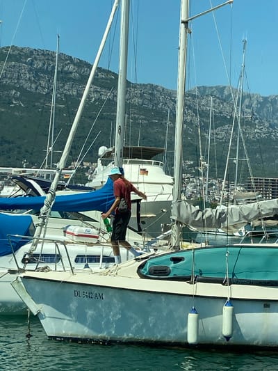 A medium shot captures a person standing on a sailboat in the marina of Bar, Montenegro. The person, wearing a red t-shirt, dark shorts, white socks, and a light blue hat, is holding a green bottle and appears to be in the process of discarding it overboard. The sailboat has the registration number "DL 5442 AM" visible on its hull. In the background, other sailboats and a larger white yacht with German flags are moored. Beyond the marina, the city of Bar is visible with buildings cascading down a hillside, leading up to a rugged, green-covered mountain range under a clear blue sky. The water in the foreground is a greenish-blue. Two white fenders with blue tops hang from the side of the sailboat.