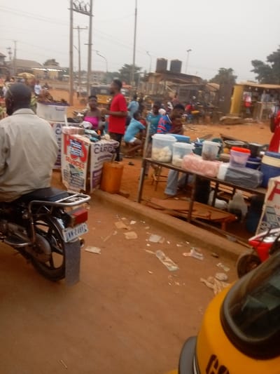 A bustling outdoor market scene unfolds under an overcast sky. People mill around vendors selling goods from tables and carts. A motorcycle sits idle on the left, its license plate visible. Litter scatters the reddish-brown ground. A yellow tricycle is in the foreground. Utilities poles rise in the background, contrasting with the low, haphazard structures housing the market. Water tanks are on a nearby building.