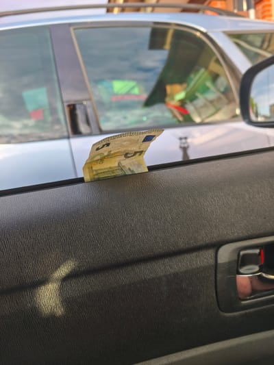 The image shows a five euro banknote tucked into the window frame of a car door. The car appears to be parked outdoors, with a reflection of buildings and trees visible in the window. The interior of the car door is made of textured black plastic, and a chrome door handle with a red accent is visible on the right side of the frame. A shadow, possibly from a nearby structure, falls across the door panel. The lighting suggests it is daytime. The banknote is the primary focus, with visible text including "EURO" and part of the European Union flag. The location is identified as Szczecin, Poland.
