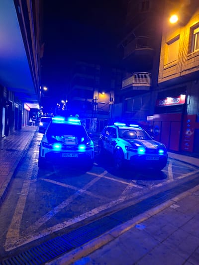 The image depicts a street scene at night in Torrevieja, Spain, illuminated by the blue flashing lights of two police vehicles. The police cars, prominently positioned in the foreground, are white SUVs with blue and yellow markings and flashing blue emergency lights on their roofs and at the front. The license plates are visible: one reads "3025 MYJ" and the other "8660 MYH". The police cars are parked in what appear to be designated parking spots marked with yellow lines on the asphalt.
To the left of the police cars is a sidewalk with a tiled surface, and beyond that, the facade of a building with shops. One shop sign, illuminated in red, reads "Foot on Mars". Further down the street, other buildings are visible, some with balconies. Streetlights cast a warm yellow glow in contrast to the blue police lights. The ground is wet, reflecting the lights. A metal grate is visible along the edge of the sidewalk in the foreground. Two individuals, appearing to be civilians, are walking on the sidewalk in the background, further down the street, illuminated by ambient light. The overall impression is a nighttime street scene where police are present, possibly on patrol or responding to an incident.