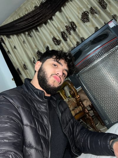 A young man with dark curly hair and a beard poses in a black puffer jacket. Behind him is a patterned curtain and what appears to be a heating unit with a grill. Part of a carved wooden object is visible in the background. The image is taken from a slightly low angle, oriented vertically. The lighting suggests an indoor setting.