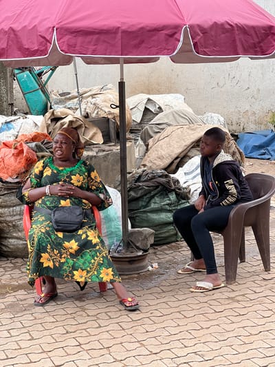 Two individuals are seated under a large, pink umbrella at an outdoor market in Nkpor, Nigeria. The person on the left is a woman wearing a green and yellow floral dress, a headscarf, and a black fanny pack. She is sitting on a red chair with her hands clasped in her lap and her mouth open as if speaking. The person on the right is a younger individual, possibly a girl, wearing a black hoodie, black leggings, and sandals adorned with pearls. She is seated on a brown plastic chair and looking to her right. The ground is paved with interlocking bricks, and the area is filled with large sacks and bundles of materials, suggesting a marketplace or storage area. A teal-colored cart is visible behind the woman.